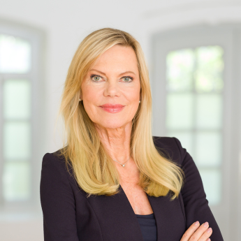 Das Bild zeigt Nina Ruge, eine blonde Frau mit langen Haaren, die selbstbewusst in die Kamera schaut. Der Hintergrund ist hell und wirkt freundlich, vermutlich ein Innenraum mit großen Fenstern.