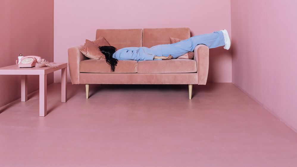 Dunkelhaarige Frau in hellblauem Anzug hat sich in einem rosa Raum auf ein rosa Sofa fallen lassen