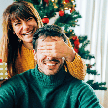 Lachende Frau hält Mann im weihnachtlich geschmückten Raum die Augen zu; Geschenk in der Hand.