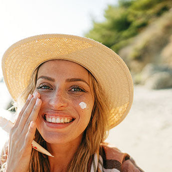 Lächelnde Frau mit Sonnenhut trägt Sonnencreme auf ihr Gesicht auf – effektiver UV-Schutz an einem Sommertag am Strand