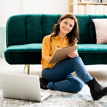 Dunkelhaarige Frau in gelber Bluse sitzt vor grünem Sofa mit Ntizblock und Stift in der Hand und einem aufgeklappten Laptop neben sich.