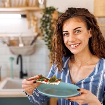 Eine junge Frau mit lockigen Haaren und einem freundlichen Lächeln hält einen Teller mit einem Knäckebrot, das mit Frischkäse, Rucola und Tomaten belegt ist. Sie steht in einer gemütlichen Küche und strahlt Zufriedenheit aus.