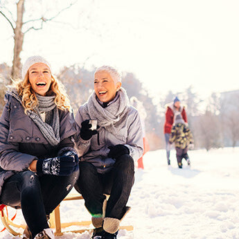 Zwei glückliche Frauen sitzen auf einem Schlitten inmitten einer Schneelandschaft.