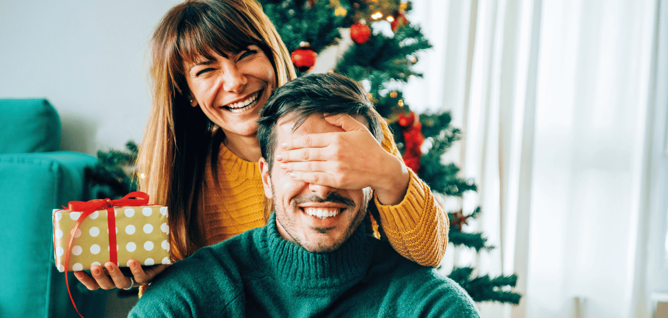 Lachende Frau hält Mann im weihnachtlich geschmückten Raum die Augen zu; Geschenk in der Hand.