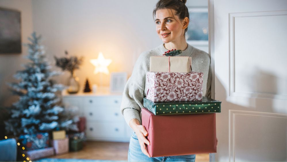 Brünette Frau im Wohnzimmer mit vier Geschenken im Arm, im Hintergrund ein Tannenbaum mit Geschenken