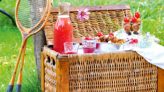 Picknickkorb auf grüner Wiese mit Spießen, Limonade befüllt und angelehnten Tennisschlägern