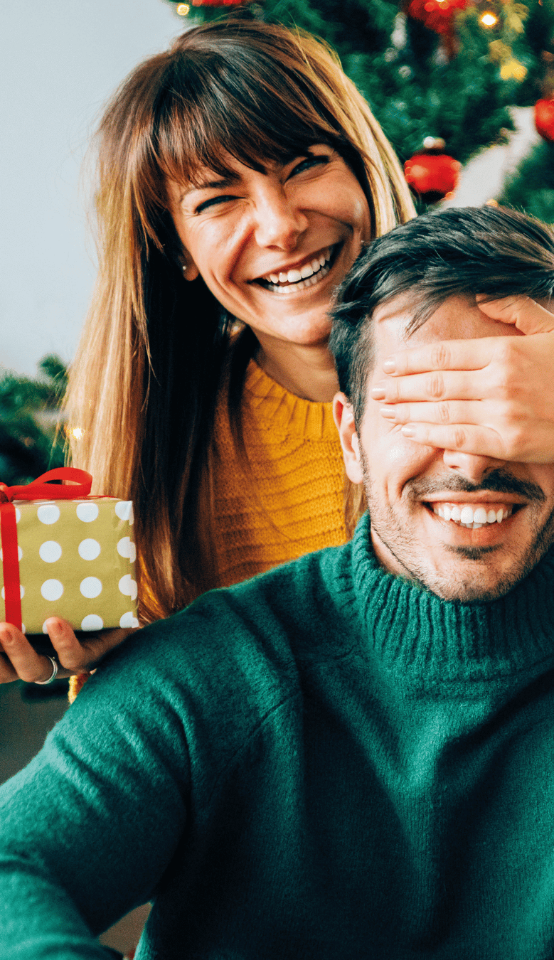Lachende Frau hält Mann im weihnachtlich geschmückten Raum die Augen zu; Geschenk in der Hand.