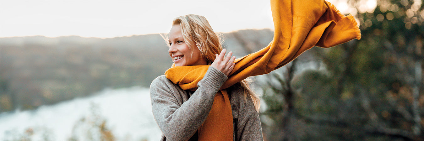 Blonde, lächelnde Frau sitzt in der Natur und schwingt ihren orangen Schal um den Hals