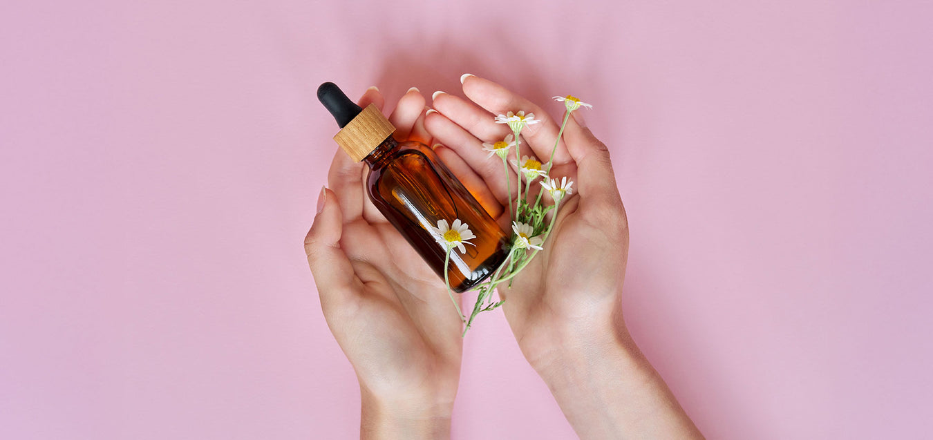 Frauenhände mit Kosmetiköl in brauner Flasche und Kamillenblumen vor rosa Hintergrund