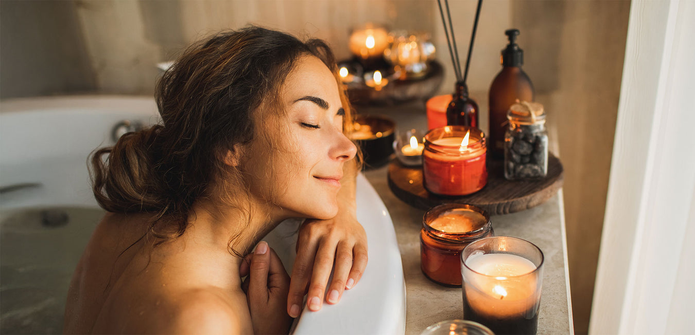 Dunkelhaarige Frau liegt entspannt mit dem Kopf auf dem Rand einer Badewanne, Kerzen leuchten um sie herum