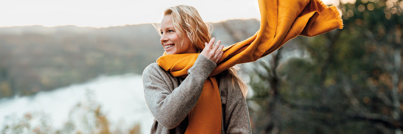 Blonde, lächelnde Frau sitzt in der Natur und schwingt ihren orangen Schal um den Hals