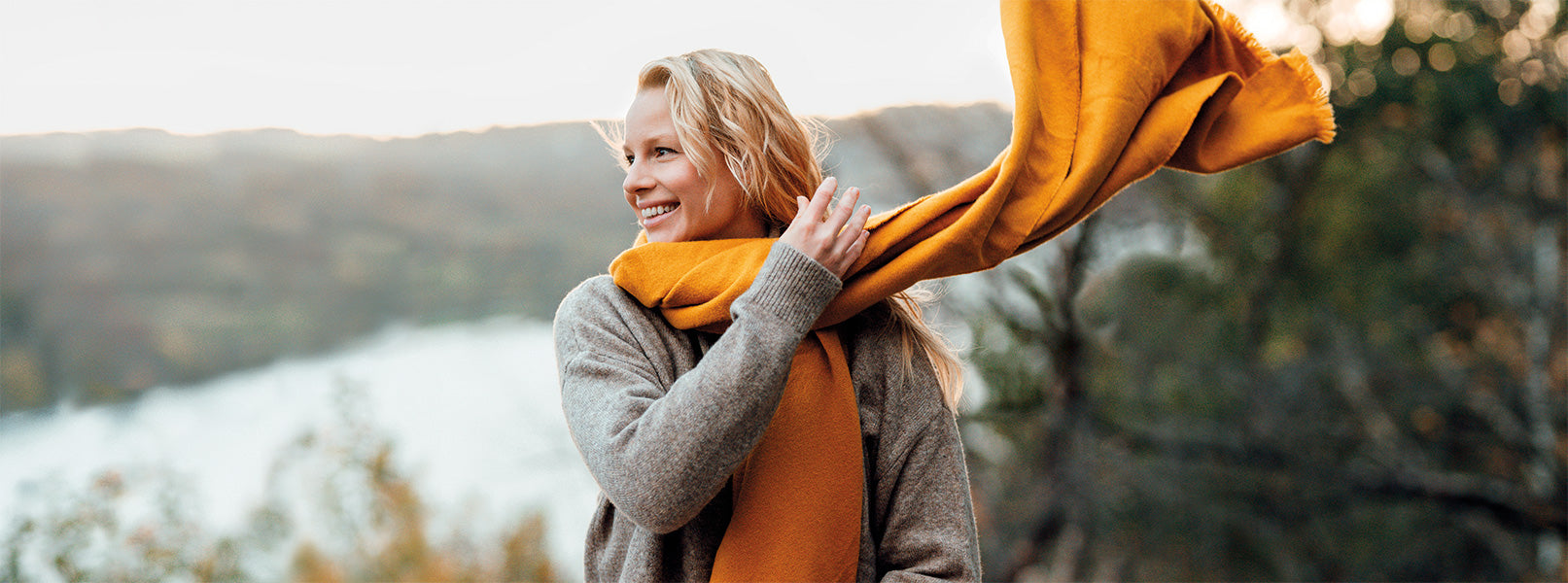 Blonde, lächelnde Frau sitzt in der Natur und schwingt ihren orangen Schal um den Hals