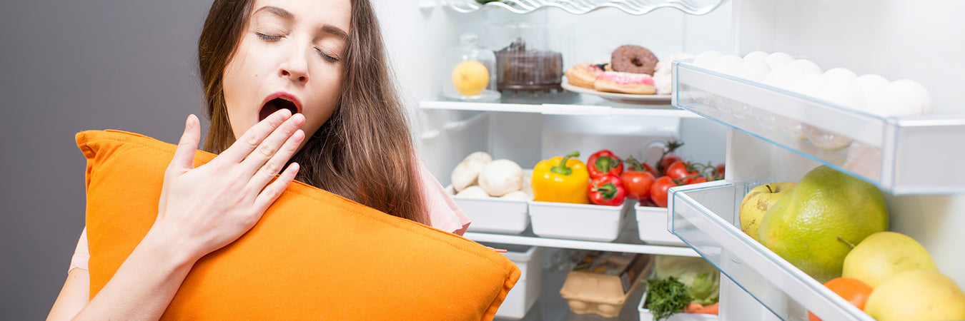 Brünette Frau steht gähnend und mit einem orangen Kissen vor der Brust vor einem geöffneten Kühlschrank