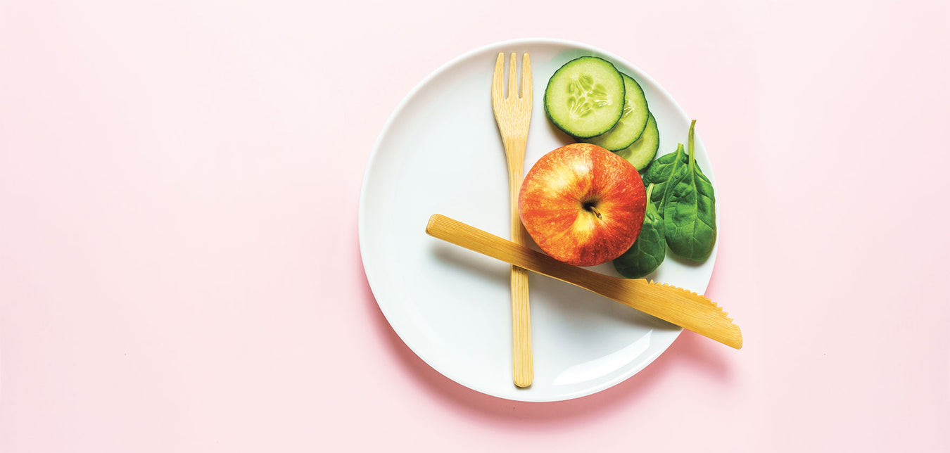 Weißer Teller mit Bambus-Besteck, einem Apfel, drei Gurkenscheiben und drei Soinatblättern steht auf einem rosé farbenem Untergrund
