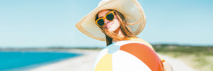 Junge, brünette Frau mit gelber Sonnenbrille, Strohhut und buntem Ball am Strand