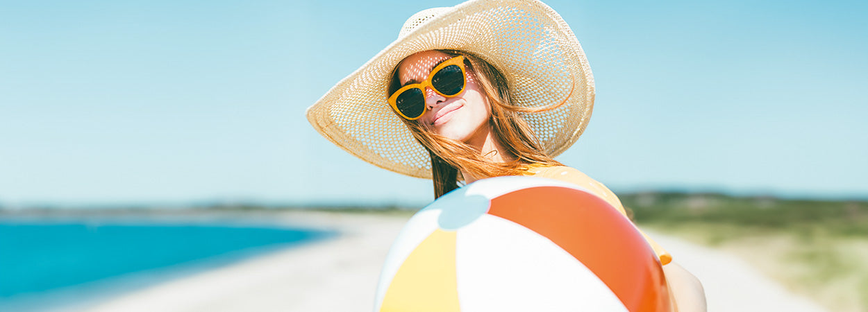 Junge, brünette Frau mit gelber Sonnenbrille, Strohhut und buntem Ball am Strand