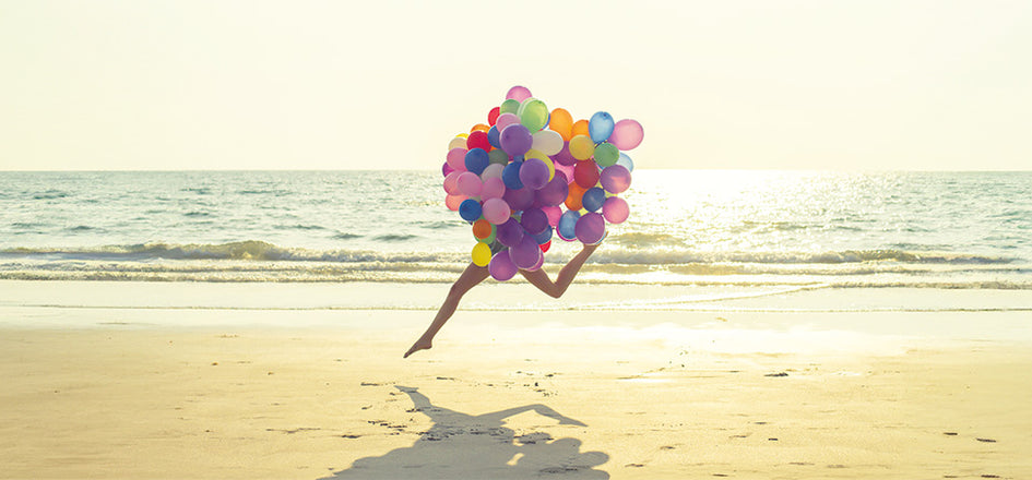 Frau, deren Oberkörper von vielen bunten Lufballons verdeckt wird, springt mit langen Beinen am Strand