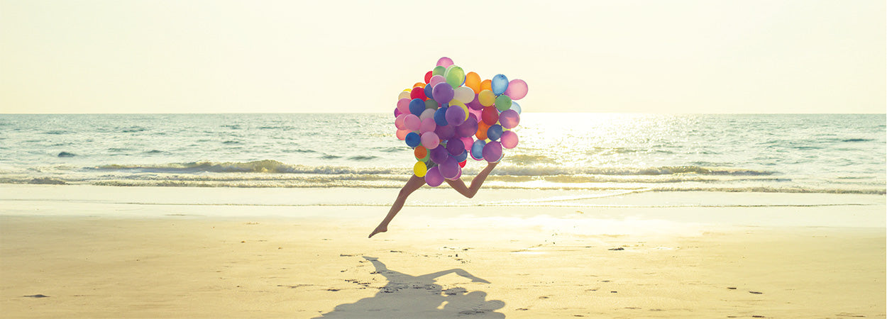 Frau, deren Oberkörper von vielen bunten Lufballons verdeckt wird, springt mit langen Beinen am Strand