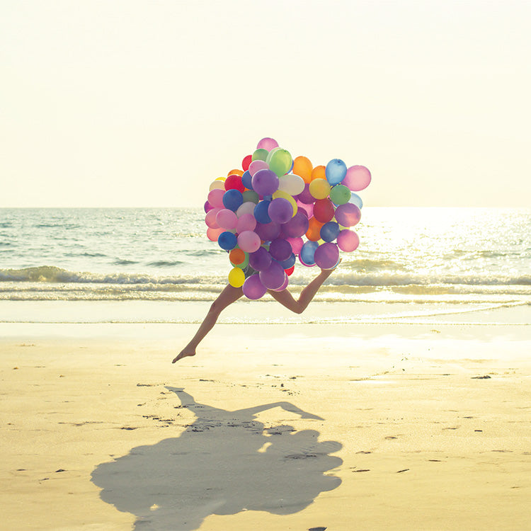 Frau, deren Oberkörper von vielen bunten Lufballons verdeckt wird, springt mit langen Beinen am Strand