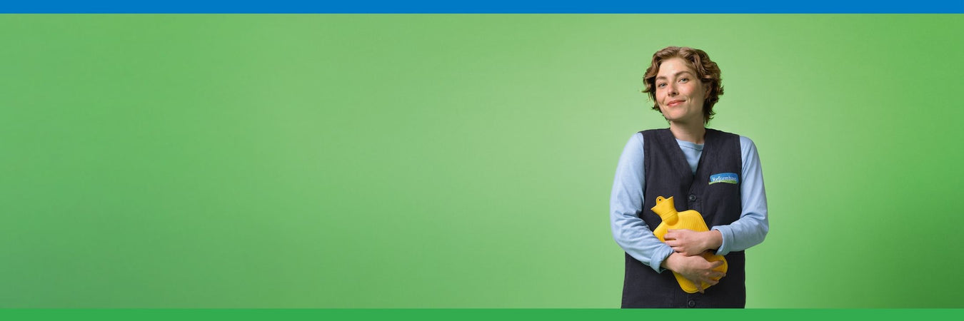 Kurzhaarige Frau im mit Reformhaus Weste und gelber Wärmflasche vor grünem Hintergrund blickt in die Kamera