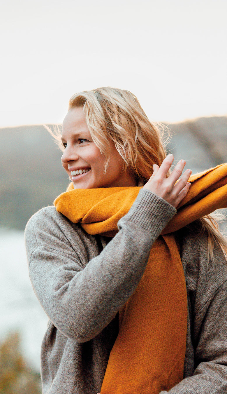 Blonde, lächelnde Frau sitzt in der Natur und schwingt ihren orangen Schal um den Hals
