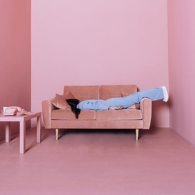 Dunkelhaarige Frau in hellblauem Anzug hat sich in einem rosa Raum auf ein rosa Sofa fallen lassen