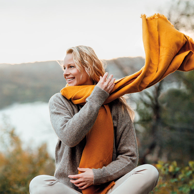 Blonde, lächelnde Frau sitzt in der Natur und schwingt ihren orangen Schal um den Hals