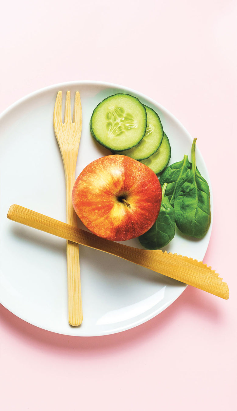 Weißer Teller mit Bambus-Besteck, einem Apfel, drei Gurkenscheiben und drei Soinatblättern steht auf einem rosé farbenem Untergrund